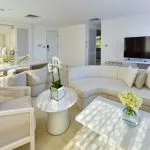 A bright, all-white luxury boat suite with a curved sofa, marble coffee tables, and a view into the bedroom.