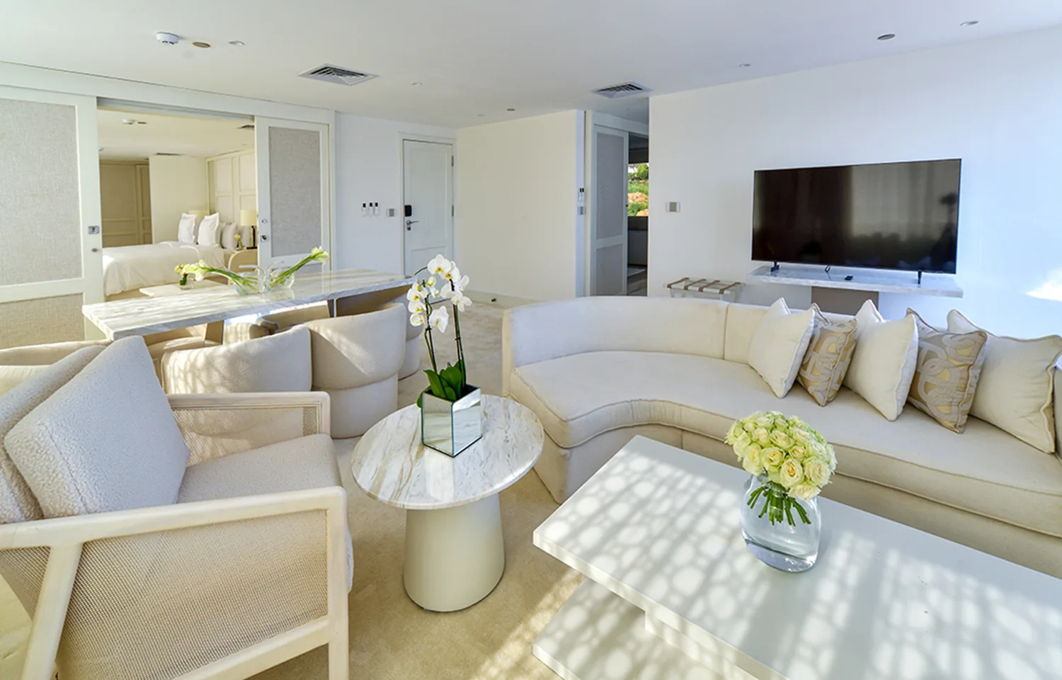 A bright, all-white luxury boat suite with a curved sofa, marble coffee tables, and a view into the bedroom.