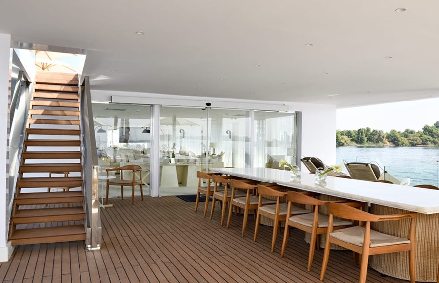 An open-air lounge on a boat deck with a long marble table, wooden chairs, and a staircase.
