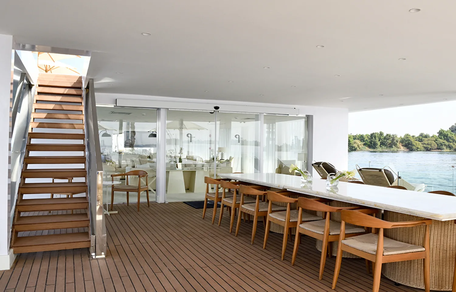 An open-air lounge on a boat deck with a long marble table, wooden chairs, and a staircase.