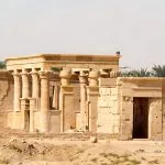 A wide shot of The Newly Renovated Hibis Temple In Kharga Egypt, showing the restored stone columns and entrance against a backdrop of desert and green palm trees.