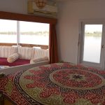 A spacious Nile cruise cabin with a double bed featuring a colorful geometric bedspread and a large window overlooking the river and an outdoor seating deck.