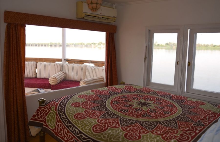 A spacious Nile cruise cabin with a double bed featuring a colorful geometric bedspread and a large window overlooking the river and an outdoor seating deck.
