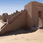 The sunlit mud-brick wall and staircase of The Roman Fortress And Temple Of Dush Baris Oasis Western Desert Of Egypt, surrounded by desert sand and clear blue sky.