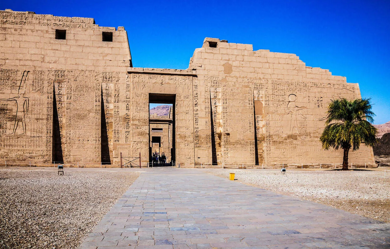 The colossal first pylon of the temple of Medinet Habu in Egypt, showing the monumental entrance and massive walls with carved reliefs against a vibrant blue sky.
