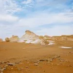 Wide landscape view of the striking white limestone formation rocks in the Western White Desert, Egypt, with a sandy foreground under a bright sky with wispy clouds.