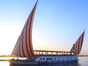 A traditional Egyptian dahabiya sailboat with red and white striped sails glides on the Nile River at sunset.