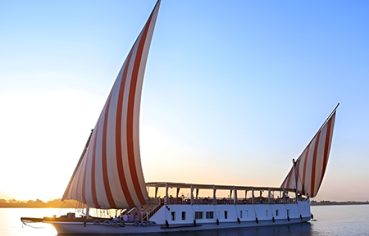 A traditional Egyptian dahabiya sailboat with red and white striped sails glides on the Nile River at sunset.