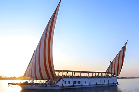 A traditional Egyptian dahabiya sailboat with red and white striped sails glides on the Nile River at sunset.