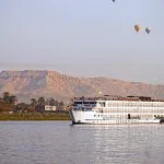 A white Nile cruise ship sailing with hot air balloons and desert mountains in the background.