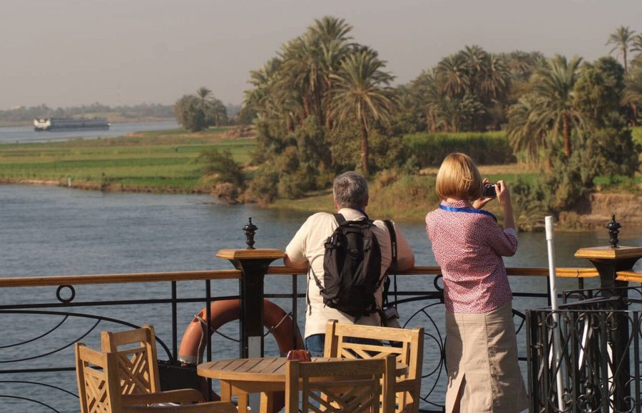 Tourists Enjoing the beauty of Egypt with a camera on the deck of a Nile River Cruise boat, overlooking palm trees and the river.