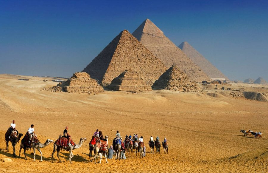 A group of Tourists riding camels at the Pyramids of Giza Cairo Egypt, with the three massive pyramids rising behind them in the sandy desert landscape.