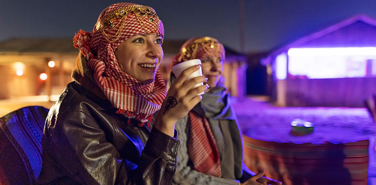 Two women tourists in keffiyehs enjoying hot drinks while watching show at Dubai desert camp