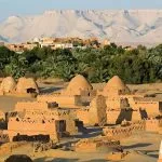 An aerial view of the mud-brick tombs and palm trees of Upper Egypt Dakhla Oasis, set against towering white and tan cliffs.