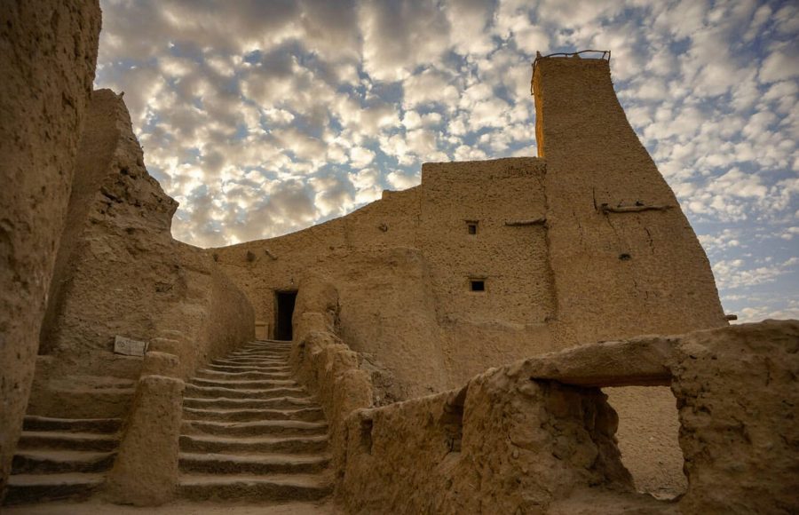 The crumbling mud-brick fortress of Shali in Siwa Oasis, with rough steps leading up to an entrance and a tall watchtower, viewed against a cloudy sky.