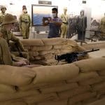 Man takes a picture of a World War II diorama featuring soldiers and a machine gun behind sandbags during a WWII battlefields tour