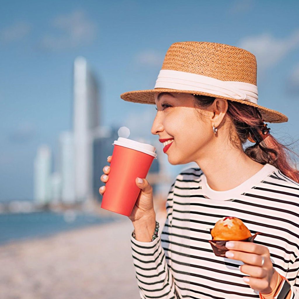 Young woman having breakfast with coffee and muffin on public beach with Abu Dhabi skyline in the background