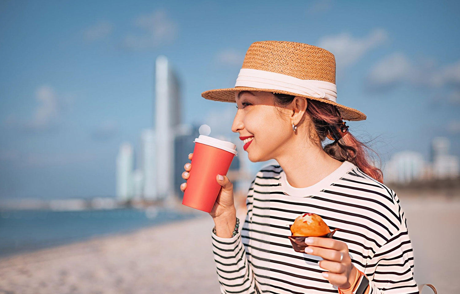Young woman having breakfast with coffee and muffin on public beach with Abu Dhabi skyline in the background