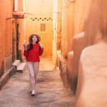 Young-woman-tourist-with-backpack-and-hat-is-walking-in-narrow-street-of-Ait-Benhaddou-Morocco-using-smartphone