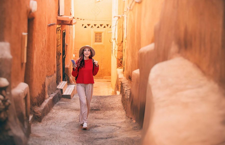 Young-woman-tourist-with-backpack-and-hat-is-walking-in-narrow-street-of-Ait-Benhaddou-Morocco-using-smartphone
