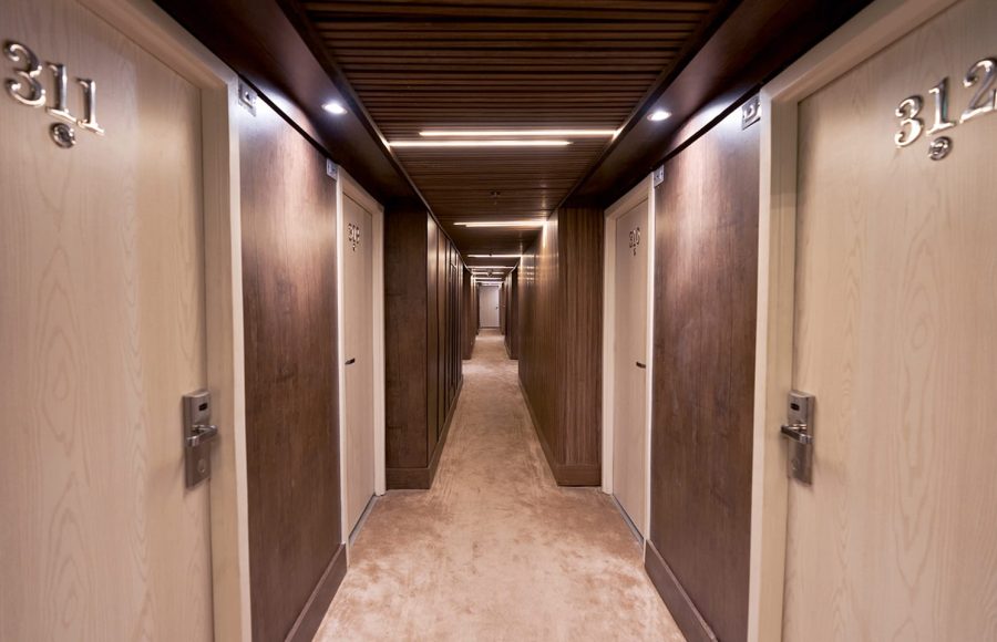 A long, modern hotel or apartment corridor with light wood doors, dark wood paneling, and a light brown carpet. The ceiling features recessed and linear LED lighting, and the doors visible in the foreground are numbered 311 and 312.