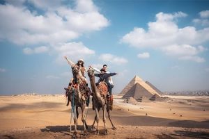 cultural-trips-in-egypt A family, likely tourists on Egypt culture trips, enjoys a camel ride in the desert with the iconic pyramids of Giza standing prominently in the background, representing Egyptian cultural heritage and a popular activity on Egypt archeological tours.