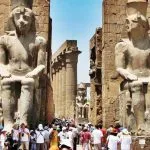 Two massive seated statues of Ramesses II flanking the entrance to Luxor Temple, with tourists walking below and columns visible in the background.
