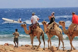 dahab-safari-day-tour Tourists ride camels along the coastline of Dahab with the Red Sea and a boat in the background, illustrating popular Dahab safari day tours and Dahab Egypt tours.