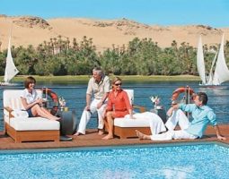 Four people relaxing by a swimming pool on a Nile River cruise ship with traditional feluccas and sandy banks in the background.