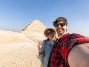 A happy tourist couple taking a selfie at Saqqara pyramid, with the ancient Saqqara Step Pyramid visible in the background against a clear blue sky.