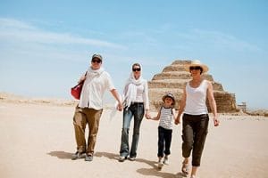 A family of four, including two adults and two children, walk hand in hand across a sandy desert landscape with the ancient Step Pyramid of Djoser visible in the background under a clear sky in Saqqara, Egypt.
