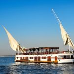 A traditional two-masted Dahabiya sailing boat on the Nile River under a clear blue sky.