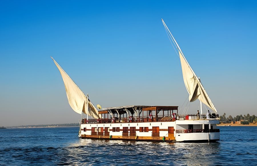 A traditional two-masted Dahabiya sailing boat on the Nile River under a clear blue sky.
