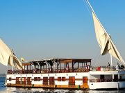 A traditional two-masted Dahabiya sailing boat on the Nile River under a clear blue sky.