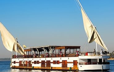 A traditional two-masted Dahabiya sailing boat on the Nile River under a clear blue sky.