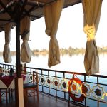 Wooden deck of a Dahabiya Nile cruise boat featuring a dining table and ornate railings overlooking the river.
