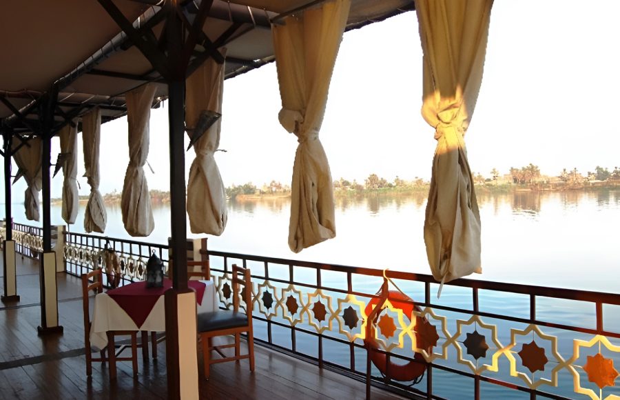 Wooden deck of a Dahabiya Nile cruise boat featuring a dining table and ornate railings overlooking the river.