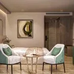 Luxury cruise cabin with a king bed, green accent chairs, light wood furniture, and a round coffee table.