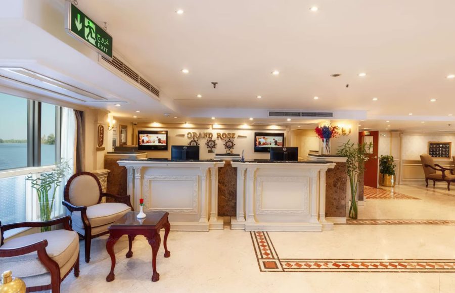 Wood-paneled cruise suite seating area with television, minibar, and queen bed visible.