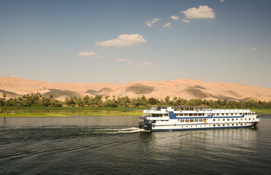 A ship sailing past the lush green banks and arid hills, representing a classic view on the Cairo to Aswan Luxor cruise.