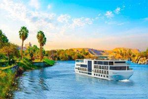 A sleek luxury cruise ship sailing along the Nile River with palm trees and a sandy landscape. Luxury Nile cruise from Aswan to Luxor.