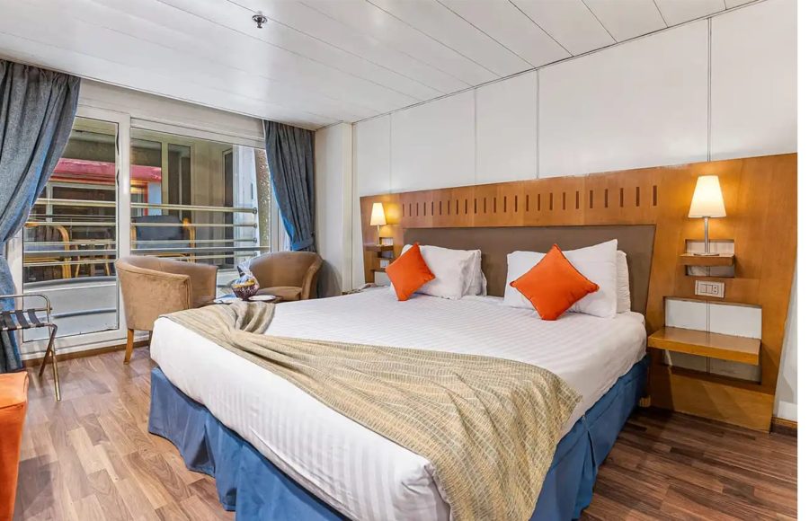 Cruise ship cabin with double bed, wooden headboard, and sliding balcony door.