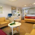 A spacious luxury suite on a cruise ship with a four-poster bed, sitting area, and elegant gold-framed wall art.