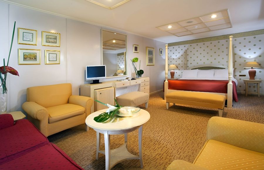 A spacious luxury suite on a cruise ship with a four-poster bed, sitting area, and elegant gold-framed wall art.