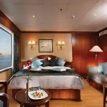 A warm, wood-paneled cruise cabin with a large picture window showing the river, a plush bed, and a sofa.