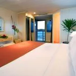 A warm, wood-paneled cruise cabin with a large picture window showing the river, a plush bed, and a sofa.