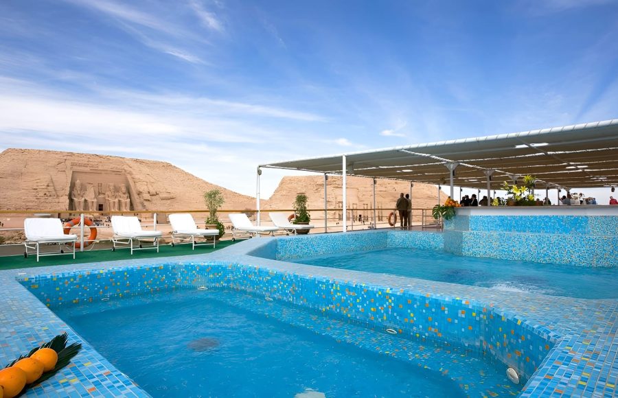 A blue-tiled swimming pool and sun loungers on a cruise ship deck with the Abu Simbel temples in the background.
