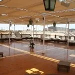 A large wooden sundeck on a cruise ship covered by a tan canopy with low seating and hanging lanterns.