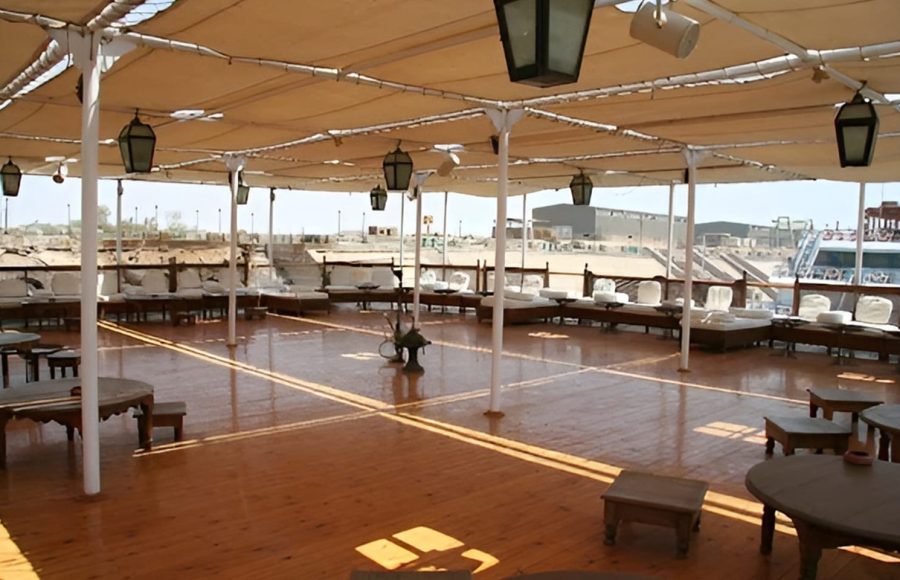 A large wooden sundeck on a cruise ship covered by a tan canopy with low seating and hanging lanterns.