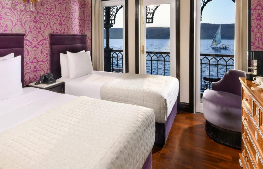A luxurious twin-bed cabin on the mövenpick ss misr steamer cruise with a balcony door open to a beautiful view of the River Nile.